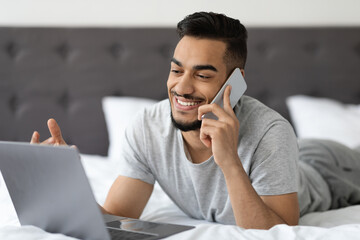 A smiling young Arab man is talking on his cellphone while using a laptop in bed. He appears relaxed and engaged, showcasing a comfortable work-from-home environment.