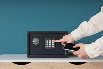 Woman with mobile phone opening safe on table near blue wall, closeup