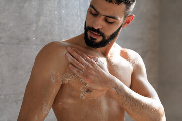 Confident young muscular man takes a refreshing shower in a modern bathroom. Soap foam covers his chest and torso as warm water cascades over him, enhancing his body care routine. © Prostock-studio