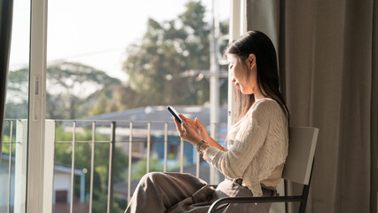 Young woman using smartphone sitting on chair at home near window