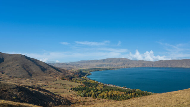 Panoramic landscape of Lake Sevan, Armenia. The deep blue water is surrounded by golden hills and green forests under a vivid clear sky.