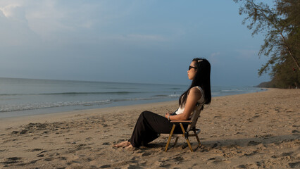 Woman sitting on a beach chair facing the sea under soft golden sunset light.