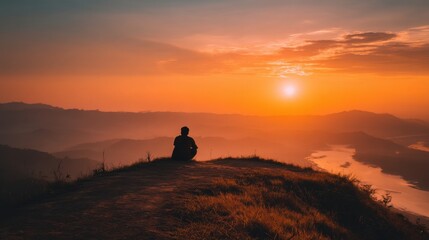 Fototapeta premium A person sits on a grassy hilltop watching the sun set over a serene river and valley. The warm colors of twilight fill the sky creating a tranquil atmosphere.