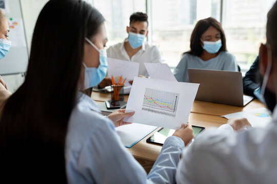 A team of employees wearing medical face masks engages in a meeting at a corporate office. One female employee is presenting financial growth data while others focus on the discussion. - Powered by Adobe