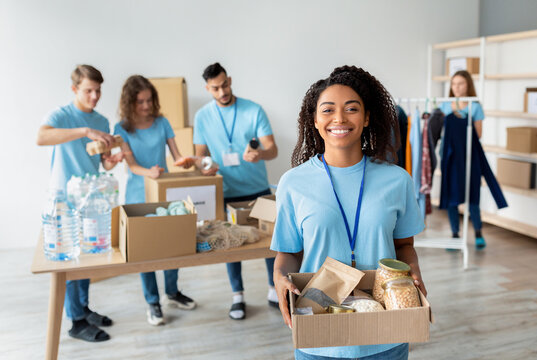 A joyful african american female volunteer in a blue uniform holds a box of food donations. In the background, other volunteers are organizing supplies in a charity center.