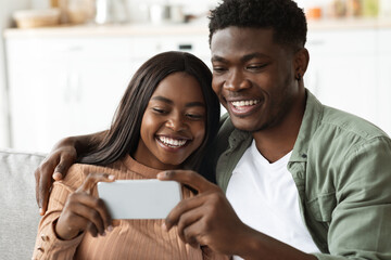 A joyful african american couple is sitting closely on their couch, smiling and embracing each other as they take a selfie together.