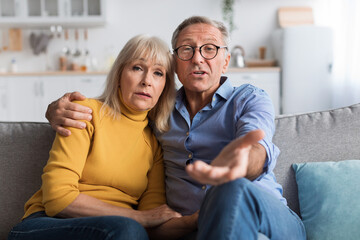 Obraz premium A middle-aged couple sits closely on a couch in their living room. They appear concerned, discussing something important. The warm room is decorated simply, creating a comforting atmosphere.
