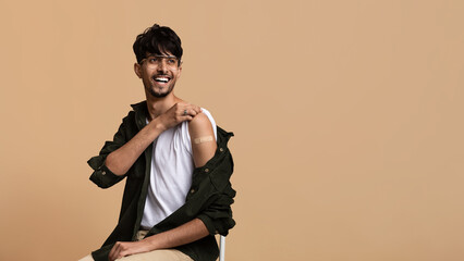 A young man sitting on a chair smiles while rolling up his sleeve to display a bandage from a recent vaccination. The background is a warm, neutral color.