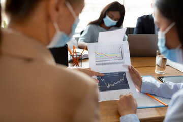 Diverse colleagues work together in a boardroom, studying financial documents and charts while wearing protective face masks. Their focus reveals a collaborative effort in a professional setting.
