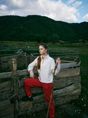 Fototapeta premium Woman on a rustic fence in a green countryside, holding a wooden staff, wearing white sweater and red pants, with hills in the background and cloudy sky.