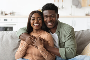 Closeup portrait of a joyful black couple embracing on a couch in their living room. They share smiles while enjoying a relaxing weekend at home, radiating love and happiness.