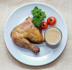 Top view of roasted chicken leg quarter served with creamy sauce, tomatoes, and parsley on white plate over rustic fabric background. Ideal for menus, recipes, and comfort food concepts