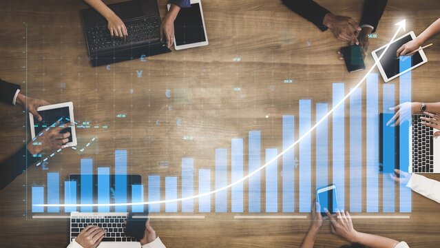 A group of professionals collaborates around a wooden table, utilizing laptops and tablets while analyzing business data through charts and graphs in a modern office setup. Copula - Powered by Adobe