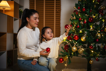 mother and toddler girl decorating Christmas tree in home at night
