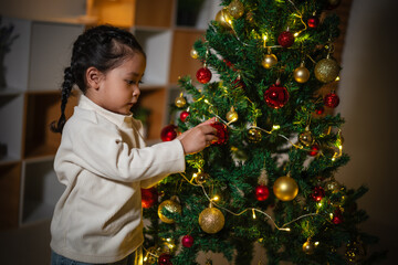 toddler girl decorating toy ball on Christmas tree in home at night