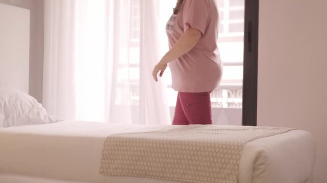 Woman 50 standing by bedroom window, making bed, dreamy look, morning light, peaceful home atmosphere, domestic serenity, simplicity morning moment, mental space, well-being, self-reflection