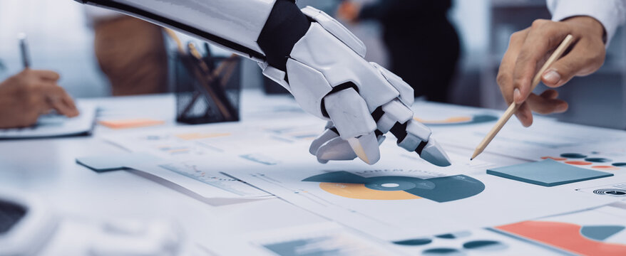 A robotic hand reaches towards business charts in a modern office setting, showcasing collaboration among diverse professionals analyzing data for informed decisions.Noogenesis - Powered by Adobe