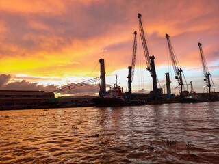 Fototapeta premium Port cranes and industrial facilities along the Magdalena River in Barranquilla, Colombia, under a dramatic pink and purple sunset sky with calm water reflections and river traffic atmosphere.