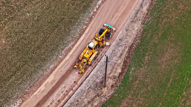 Drone follows a yellow agricultural trencher digging a narrow trench along a rural dirt road, filmed in downward angle with surrounding fields.