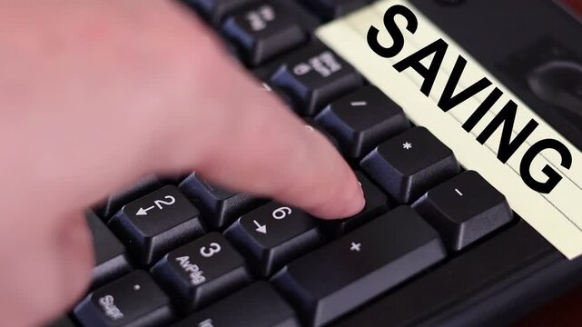 Un primer plano de un teclado con n&uacute;meros con el dedo de una mujer adulta marcando los n&uacute;meros y una nota amarilla con la palabra ahorro 