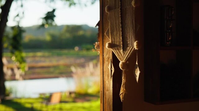 Warm light illuminates a handcrafted macrame hanging against a blurred background of a peaceful outdoor scene, featuring nature and a calm body of water.