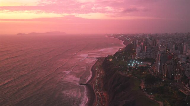 Afterglow sunset Miraflores coastal seaside aerial drone harbor Lima Peru cars traffic Makaha Barranco beach pink purple clear skies hazy cityscape skyscraper buildings ocean waves backwards up
