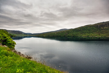 2023-06-06 A LOCH IN THE AREA OF LAIRG SCOTLAND WITH CALM WATERS AND ANICE REFLECTION AND LUSH GREENERY ON THE BANKS AND A CLOUDY SKY