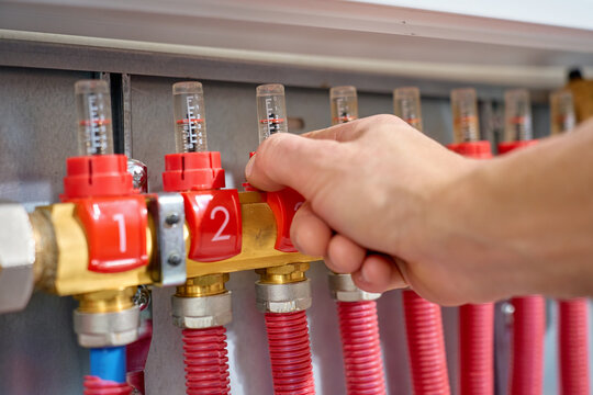 Close up of hand adjusting valve on heating manifold with flow meters and red pipes. Concept of heating system control, plumbing installation and maintenance service.