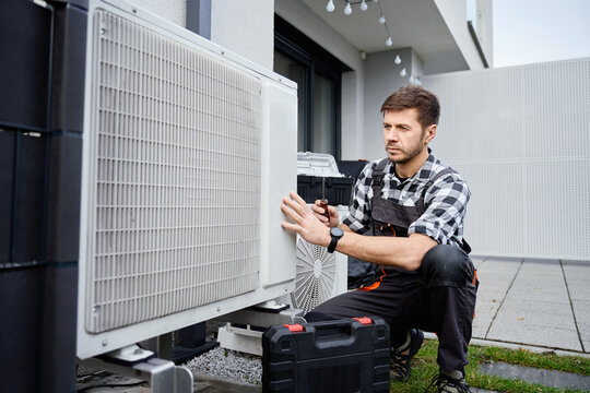 Technician performing maintenance on outdoor heat pump unit near residential building exterior. Man installing air source heat pump, using tools. Concept of HVAC service and heating system repair