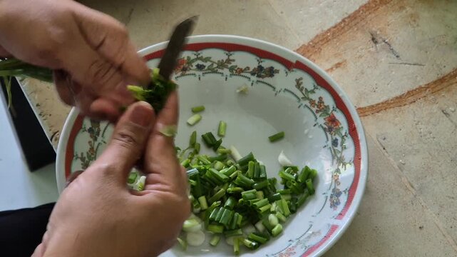 Close up hands chopping fresh green onions or scallions with a knife into a bowl for cooking ingredients.