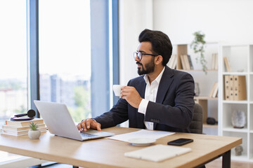 Obraz premium An Arab businessman enjoys a coffee break while working on his laptop in a modern office setting