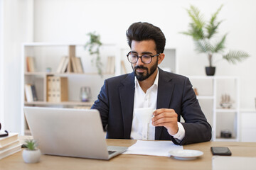 Obraz premium An Arabian businessman is working on his laptop while drinking coffee in his office