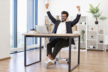 An Indian businessman is celebrating a success in his modern office, raising his arms in triumph