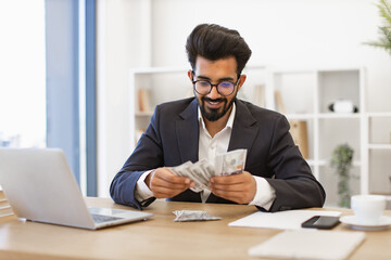An Indian businessman is smiling while counting a stack of US dollar bills at his desk