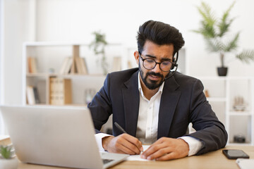 An Indian businessman wearing a headset takes notes while working on his laptop at his desk