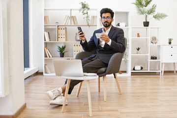 A stylish businessman is relaxing in a modern office setting, holding a cup of coffee and his phone