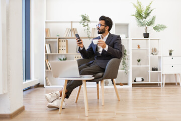 An Arab businessman is relaxing in a modern office setting, holding a phone and a cup of coffee