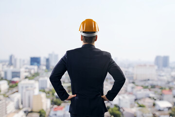 Construction businessman standing with hands on hips looking at city skyline from rooftop, rear view on bright day, concept of urban development.