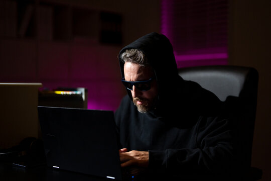 hacker types on a laptop in a dark office. Anonymous hacker in a hoodie hacking data late at night. Cyber criminal and stolen digital information concept. Hacker using laptop in night office. - Powered by Adobe