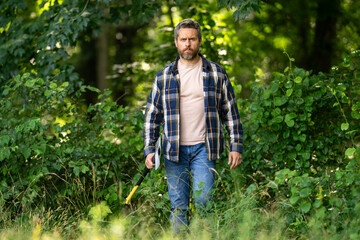 Lumberjack in the woods with an axe. Bearded man in hat with a hatchet. Lumberjack brutal man with beard and moustache in forest. Canadian lumberjack with axe. Cutting wood. Logger tools.