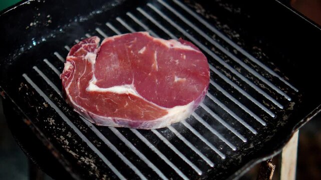 4K raw beef steak is placed on hot grill pan beginning the cooking process with visible searing marks, preparation of grilled meat dish.