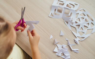 A child creating paper white snowflakes at a table at home. A child cuts patterns on paper with scissors. Decoration for the winter holiday.