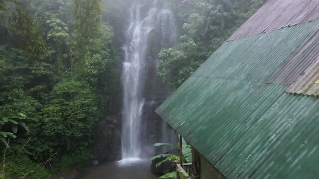 Drone view showing tall waterfall cascading beside a green roof structure in a lush forest near Gitgit Falls in Bali capturing flowing water, misty air and natural landscape in a quiet setting.