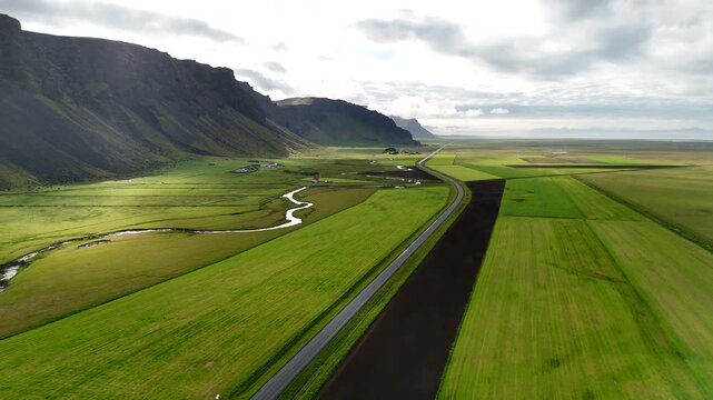 Aerial view of vibrant green fields stretching below steep moss-covered cliffs in Su&eth;urland, Iceland, with winding streams and dramatic shadows shaping the vast volcanic landscape.