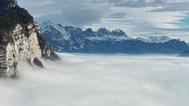 Majestic aerial view of the Churfirsten mountains and Amden, Switzerland, with rugged, snow-dusted peaks dramatically rising above a thick cloud inversion. Sunny, high-alpine scenery.