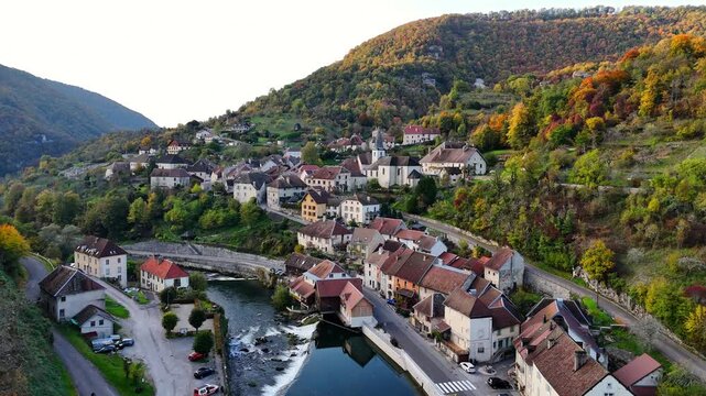 Gliding over Lods built along a bend of Loue River, with stone houses, small weir and riverside road at bottom of steep, wooded valley slopes in the Doubs department of Bourgogne‑Franche‑Comt&eacute;, France