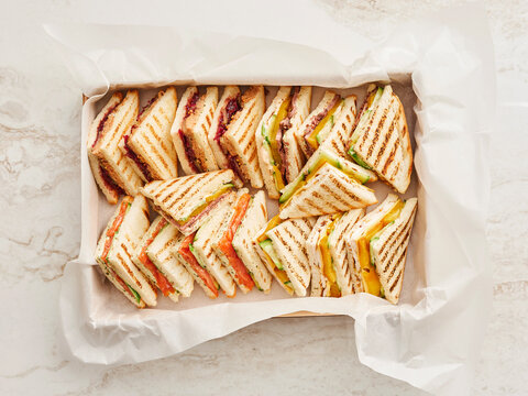 A variety of freshly made triangular grilled sandwiches are neatly arranged in a box with paper lining. Different fillings are visible, including jam, smoked salmon, and vegetables
