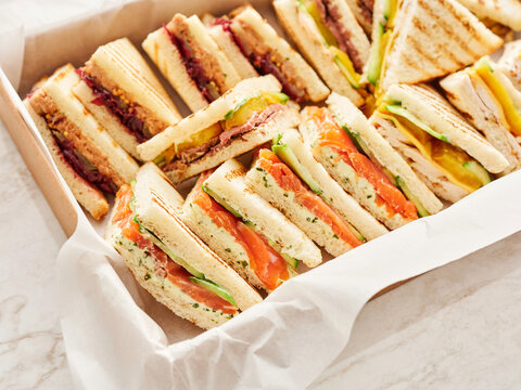A variety of triangular cut sandwiches are arranged in a box. Fillings include salmon, cucumber, chicken, cheese, beef, and jam with peanut butter
