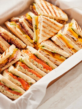 A box holds several triangle-cut sandwiches with different fillings. Fillings include salmon, ham, cheese, and other assorted ingredients. The sandwiches are arranged neatly in the box