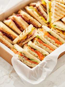 A box is filled with various sandwiches, cut into triangle shapes. The sandwiches have different fillings, including salmon, meat, and colorful vegetables. The box is on a white surface